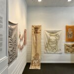 A photo of AANM gallery with textile paintings hung on the wall. they are in various organic beige and peach shades, each rippled sheet showing twisted cartoony bodies with long limbs tangled and undulating.