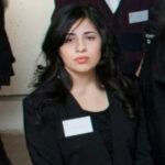 Headshot of Hope Flynn, a young light-skinned woman with shoulder-length, glossy black hair. She is wearing a simple black suit, with a name tag on the lapel, and looks towards us with a level, serious expression.