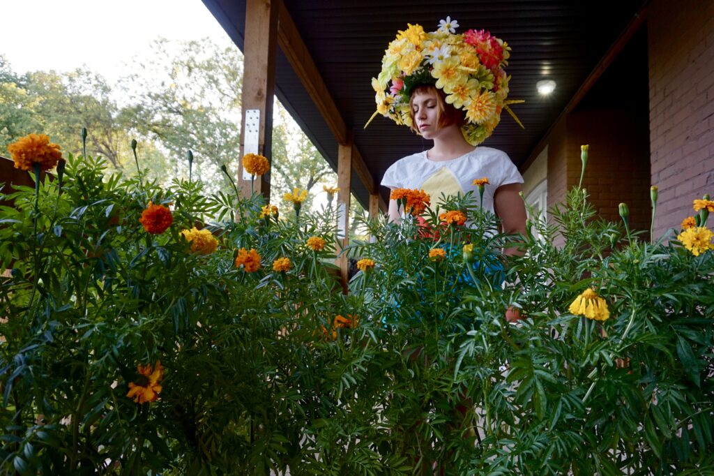 This is a photo of Katrina Craig. In the foreground is a bush of orange and yellow flowers with green leaves. Katrina is standing behind the bush and in front of a brick porch of a building. Katrina is a young white woman with chin length brown red hair with bangs. She is wearing a white t-shirt and a beautiful headpiece made up of many flowers. Her eyes are closed and her face is serene.