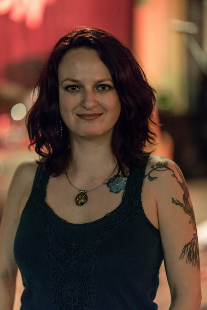 This is a headshot of Lindsey White. Lindsey is a white woman with dark auburn hair that brushes her shoulders. Her green eyes are smiling and looking directly at the camera. She is wearing a round, tigers eye pendant around her neck and a dark green tank top which shows off her nature inspired tattoos on her left shoulder, arm and collarbone.