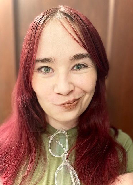 This headshot is of Hailley Rhoda. Hailley is a young white woman with long red hair. She is wearing a green t-shirt with clear framed glasses hanging from the neck of her shirt. Her elfin face is smiling a crooked smile in a mischievous grin.