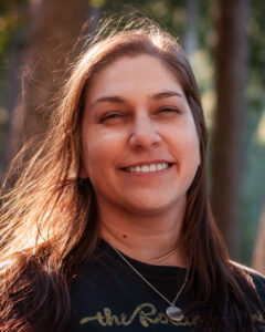 This is a headshot of Jenel Shaw. Jenel is a white woman in her 40s. She has long straight brown hair and blue eyes. She is wearing a black t-shirt and a silver necklace. She is standing outside with the sunlight in her hair smiling last the camera with squinting eyes