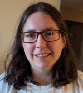 This is a headshot of Sara Arenson. Sara is a white woman in her 30s with brown eyes and brown shoulder length straight hair. Sars is wear a white t-shirt and red rimmed glasses. She is smiling and looking directly into the camera.