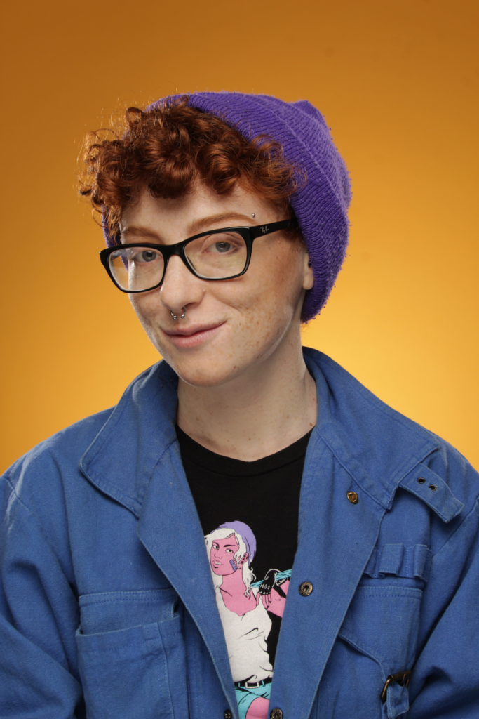 This is a photo of Kay Kassirer, a young white person with curly red hair and dark-rimmed glasses. They look at us with a mischievous smile. They sport nose jewellery, a purple knit cap, and are wearing a blue jumper over a black shirt with a drawn image of a tough looking girl with pink skin, white hair and tank, and black bike gloves. The background of the headshot is bright yellow fading to orange at the top.