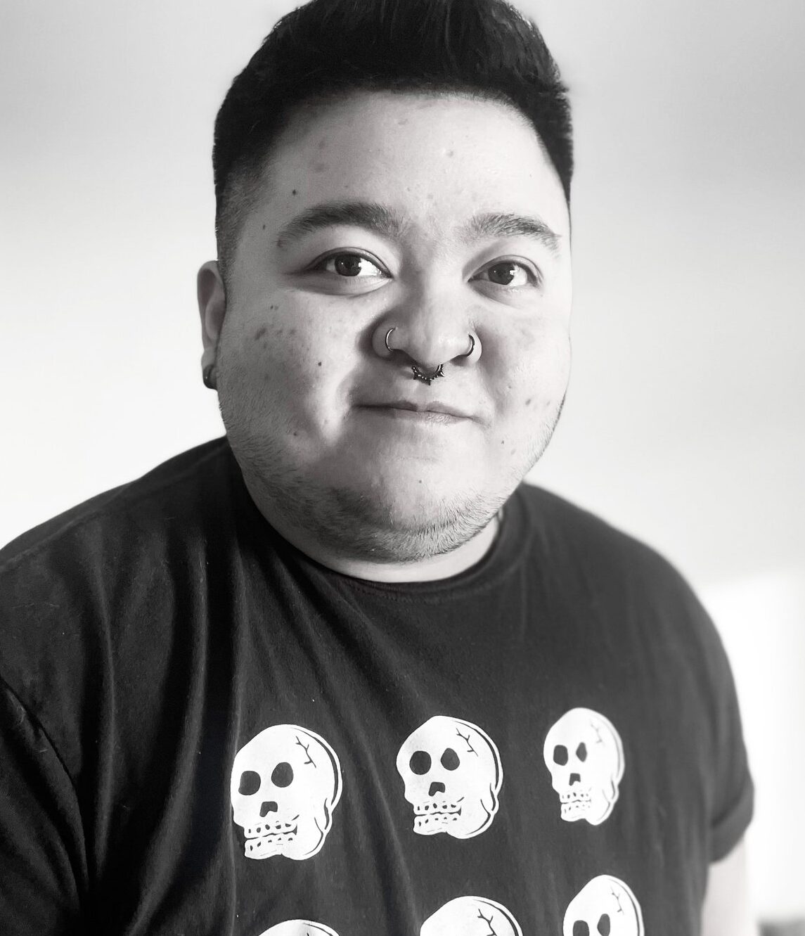 This is a photo of Nix Cristobal Calma, AANM'S Communications and Volunteer Coordinator. They are a racialized person with dark hair, wearing a black shirt with white illustrations. They are looking at the camera with a slight smile.
