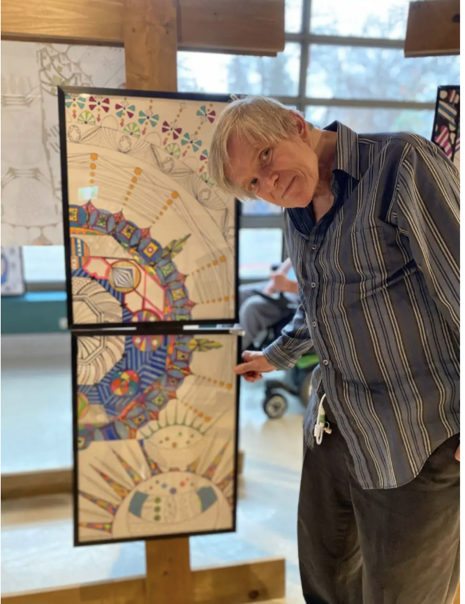 This is a photograph of Andre Grusska standing infront of his artwork. Andre is a older white man with short grey hair