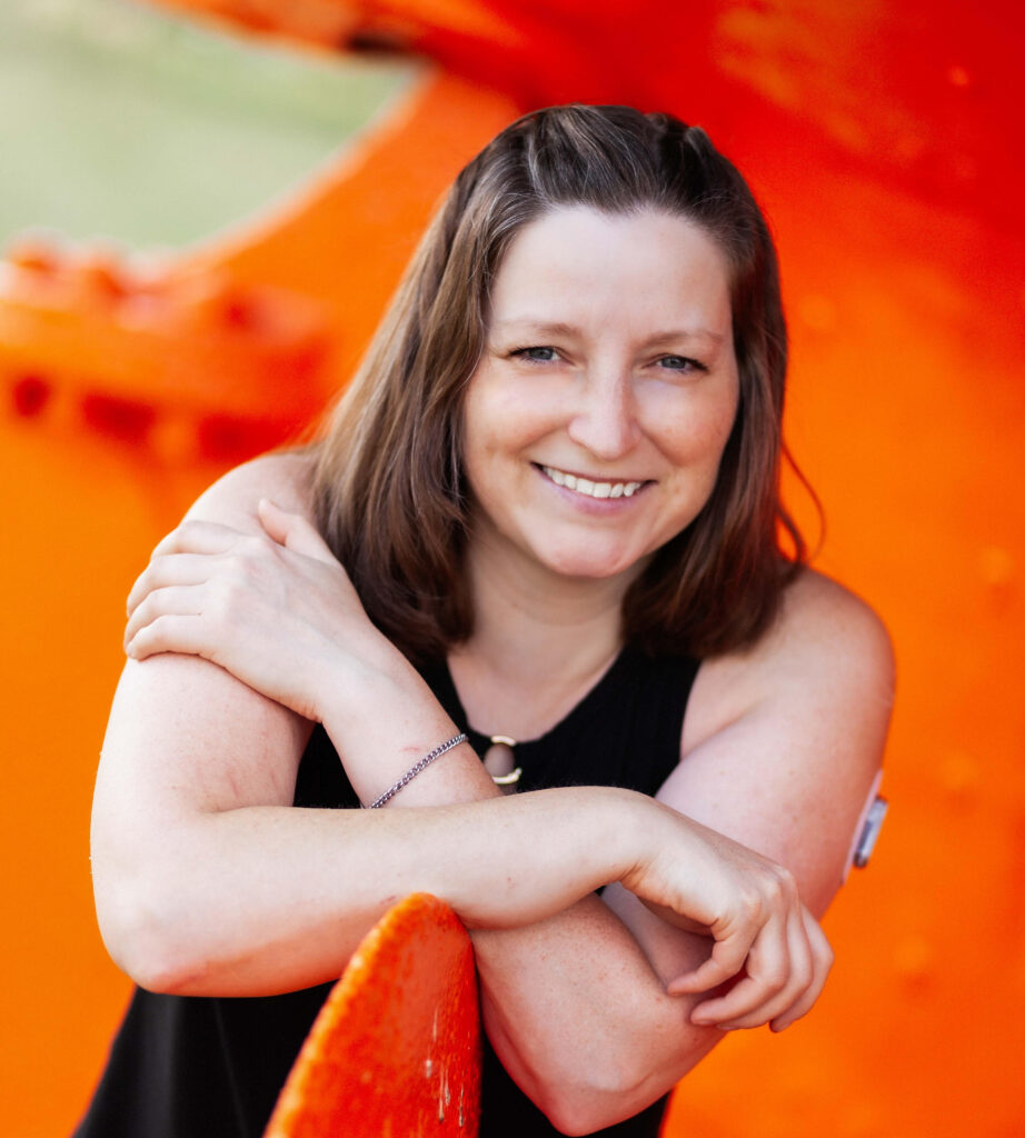 A photo of Jordan Miller. Jordan is set against an orange backdrop. She has short brown hair. She is smiling at the camera with both arms across her chest.